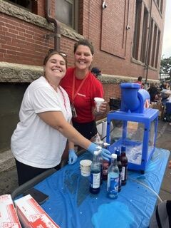 Two smiling young women in red and white shirts prepare drinks at an outdoor stand with a shaved ice machine.