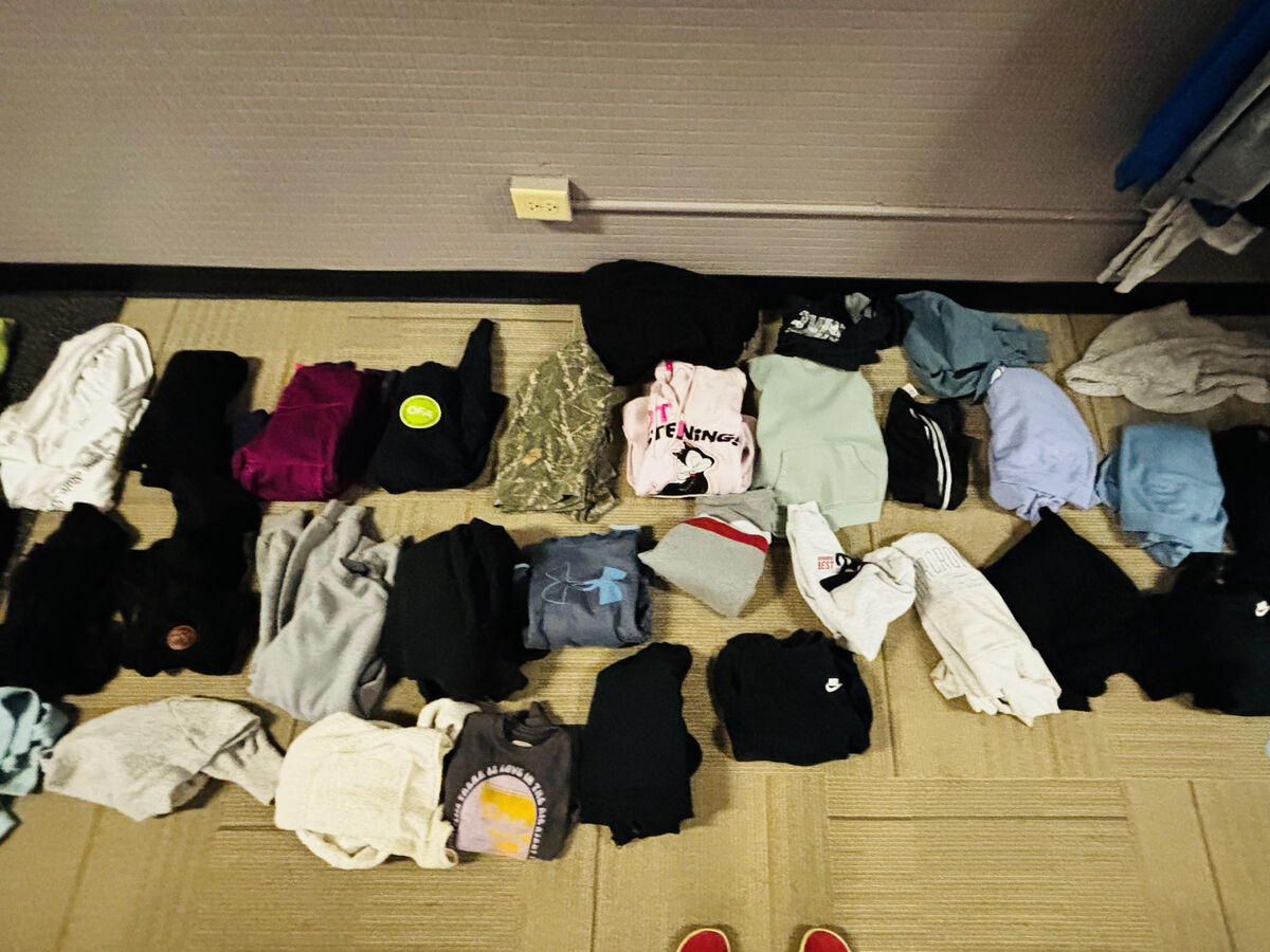 A collection of folded clothing, including hoodies, t-shirts, and pants, is laid out on a carpeted floor.
