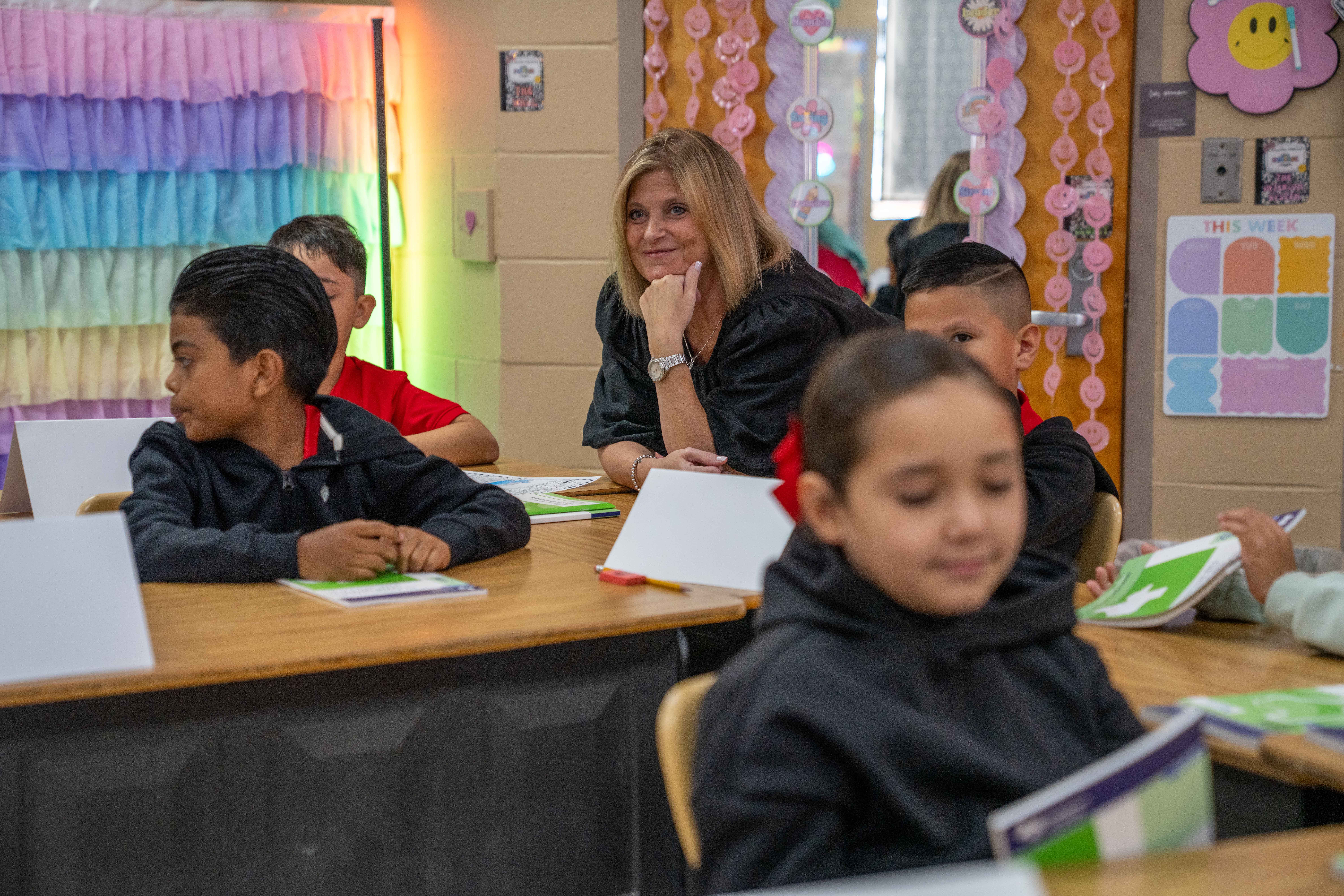 A teacher smiles while observing students at their desks in a classroom.