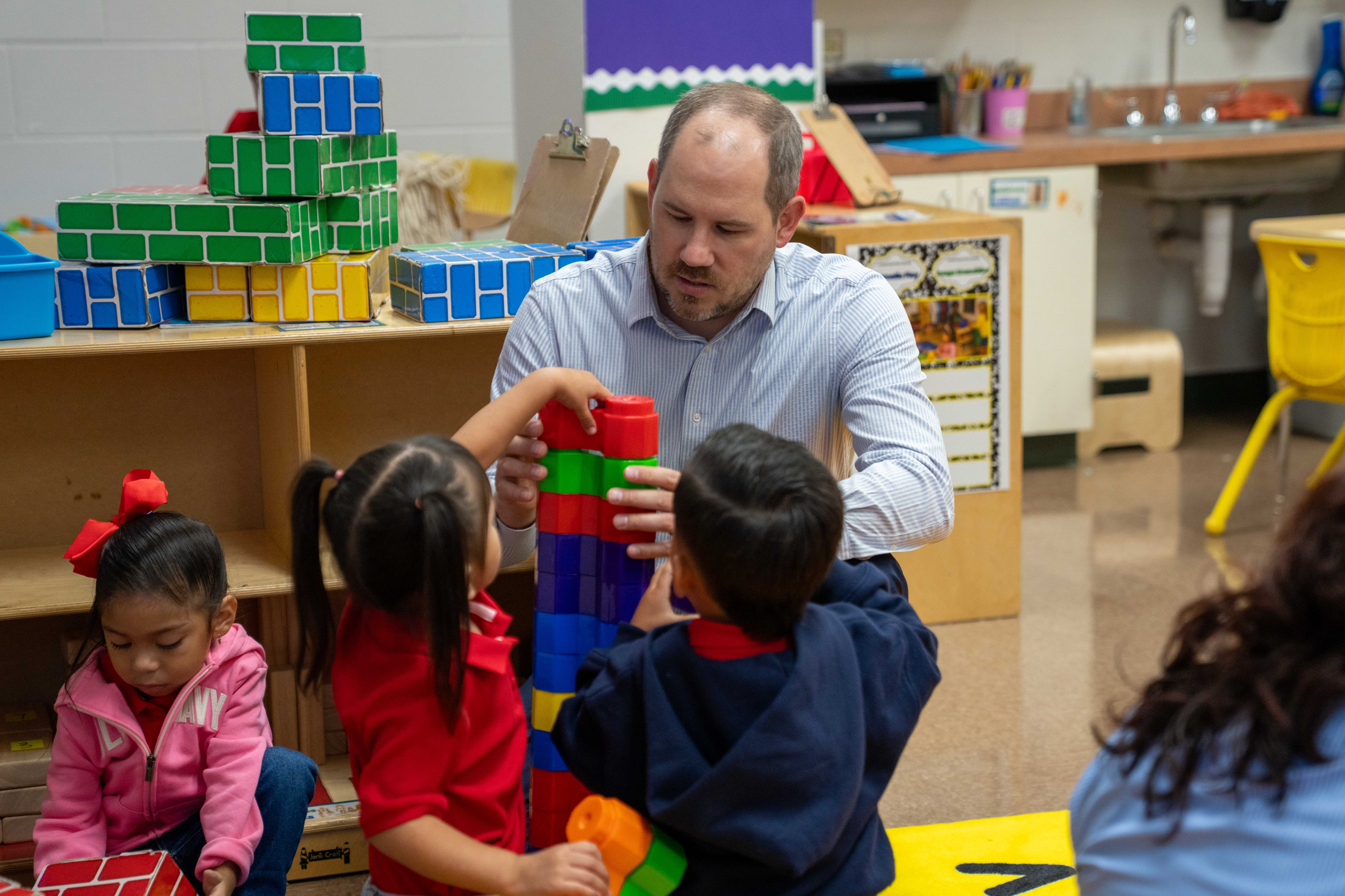 A teacher and three young children build a tall tower with colorful blocks in a classroom.