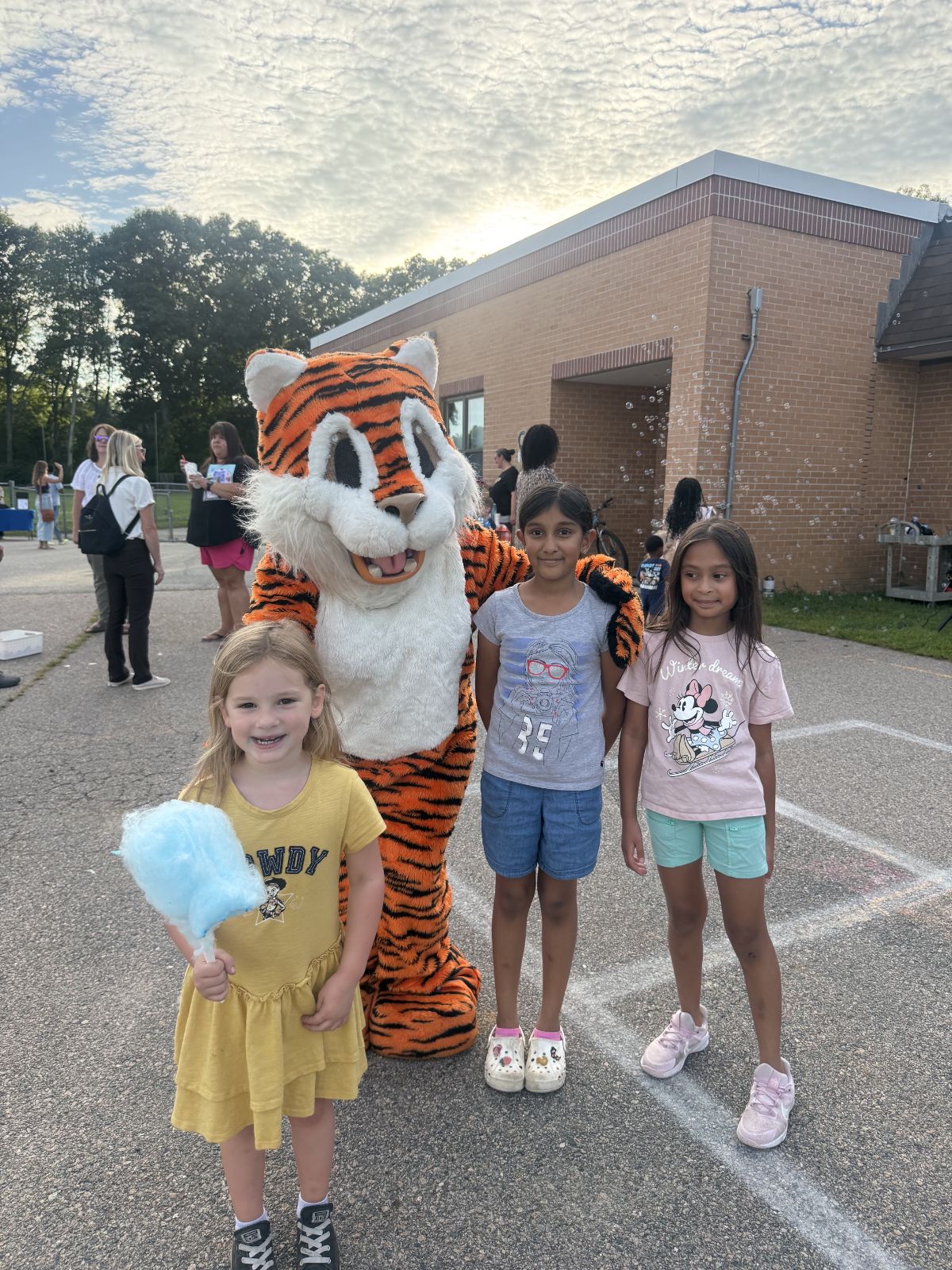 Three smiling children pose for a photo with a tiger mascot outdoors near a brick building.