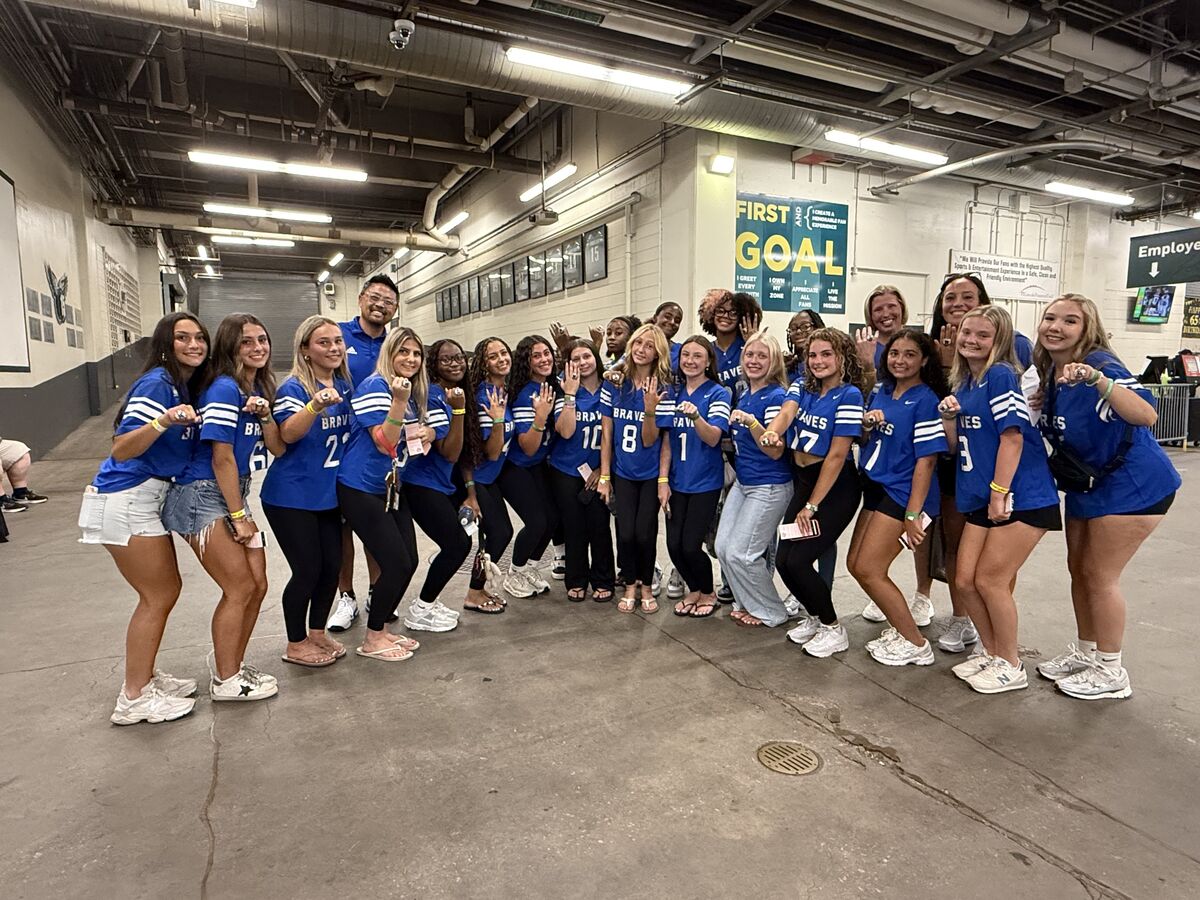 A group of young women in blue 'BRAVES' jerseys pose for a photo, many wearing championship rings.