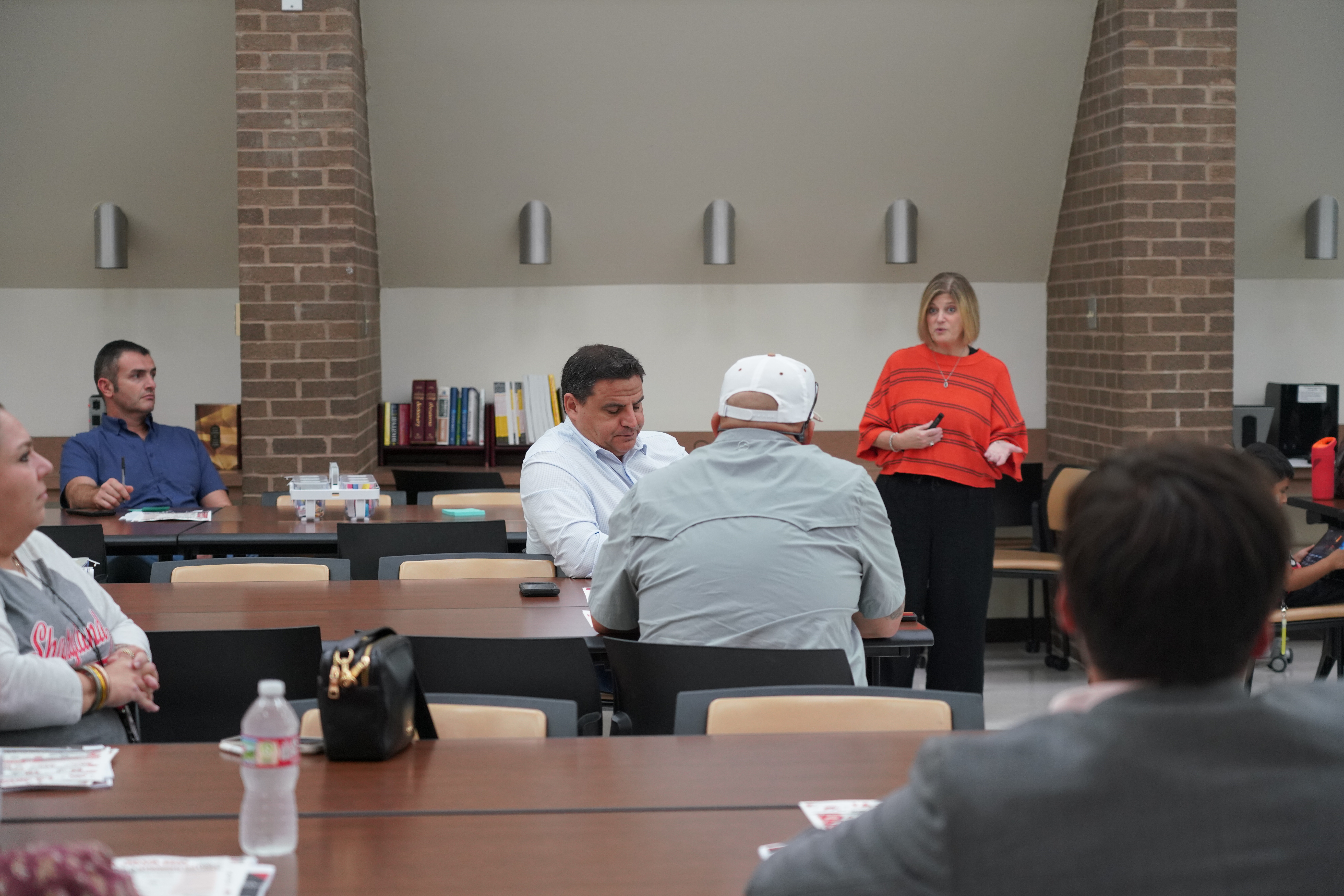 A woman in a red sweater presents to a group of people seated at long tables in a meeting room.