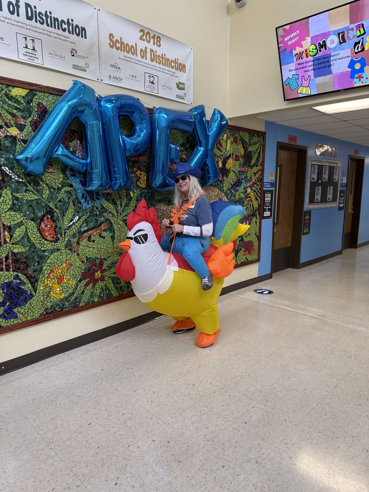 A person in an inflatable chicken costume poses with large blue '2019' balloons in a school hallway.
