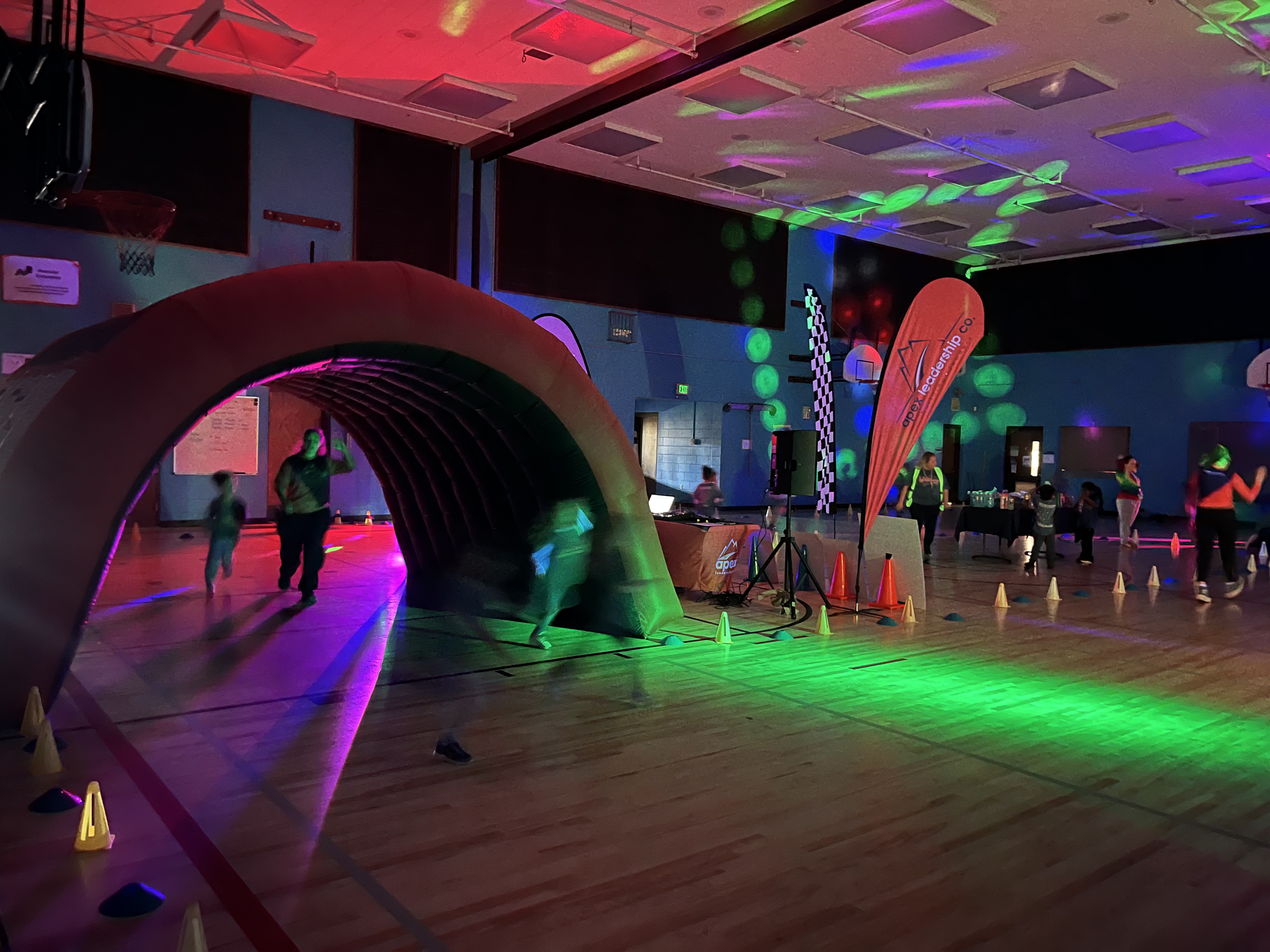 Children run through an inflatable tunnel in a gym lit with colorful disco lights.