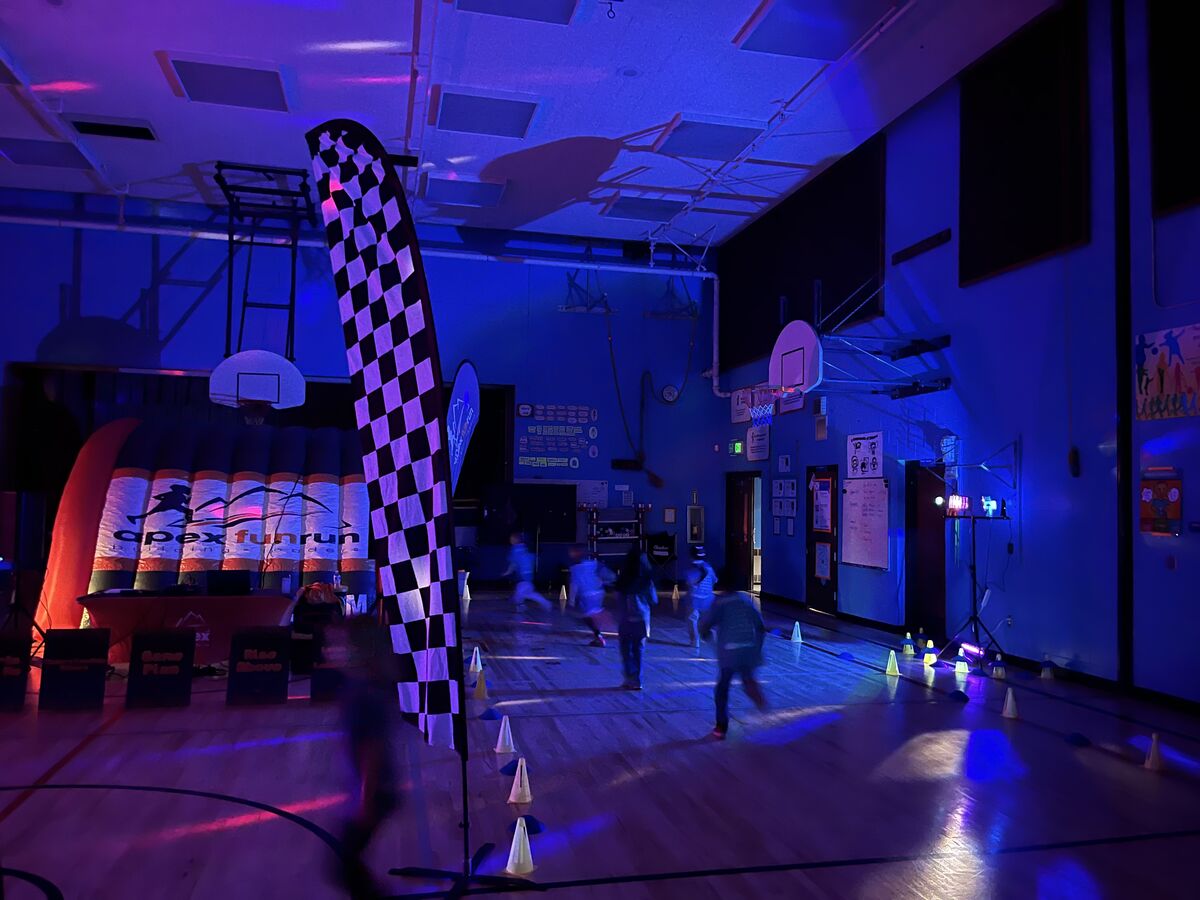 Children run through a dimly lit gymnasium decorated with a checkered flag and inflatable obstacles for an 'Apex Fun Run'.
