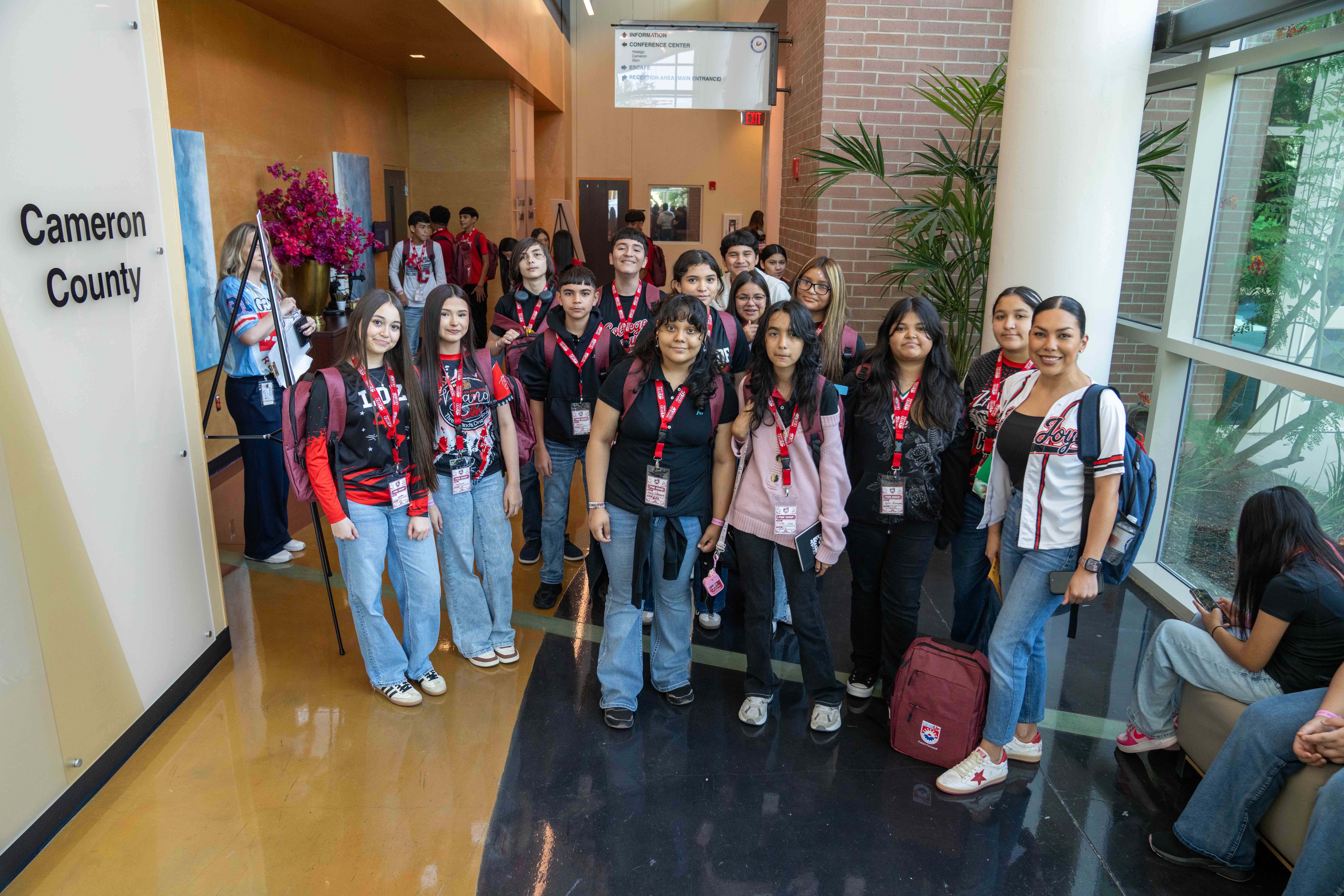 A diverse group of students and a leader pose for a photo in a brightly lit building lobby, wearing lanyards.