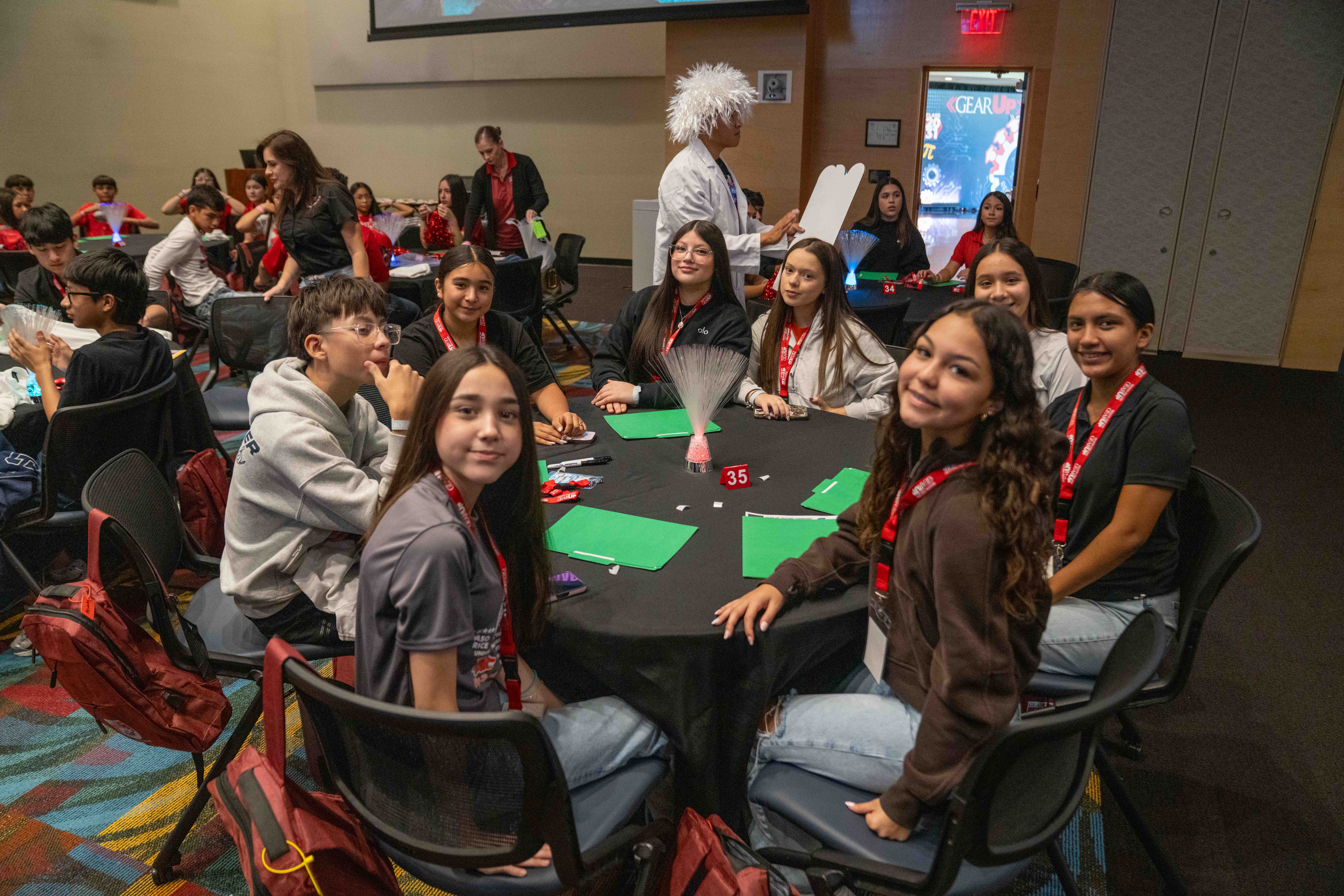 A group of students sits around a circular black table at an event, smiling and looking toward the camera.