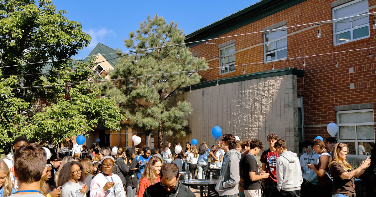 A diverse group of young people gather outdoors near a brick building, with blue and white balloons floating in the air.