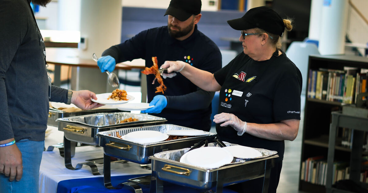 Two people in black shirts and caps serve food from chafing dishes to a person holding a plate.