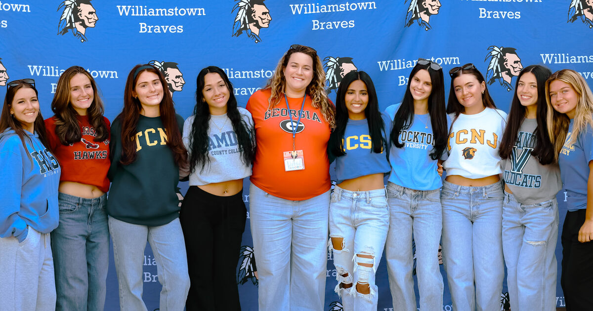 A group of young women stand together in front of a blue banner with 'Williamstown Braves' text and graphics.