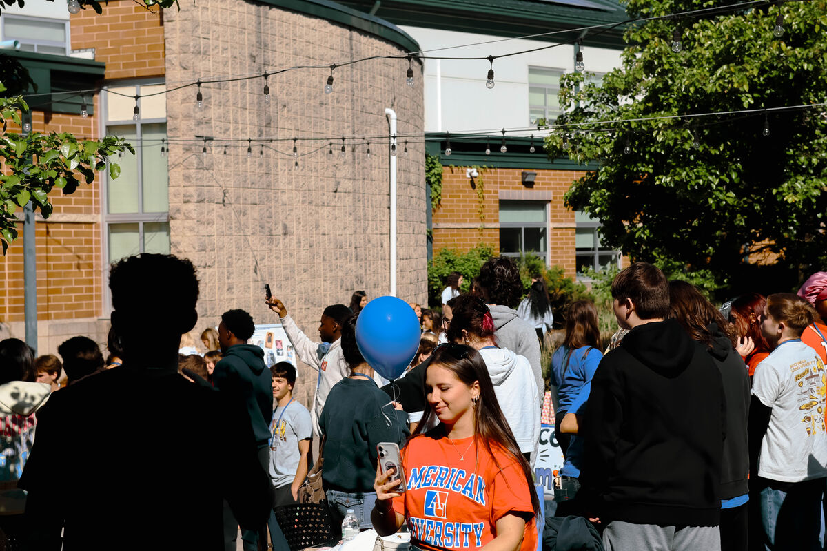 A group of young people gather outdoors, with one person holding a blue balloon and another taking a photo with their phone.