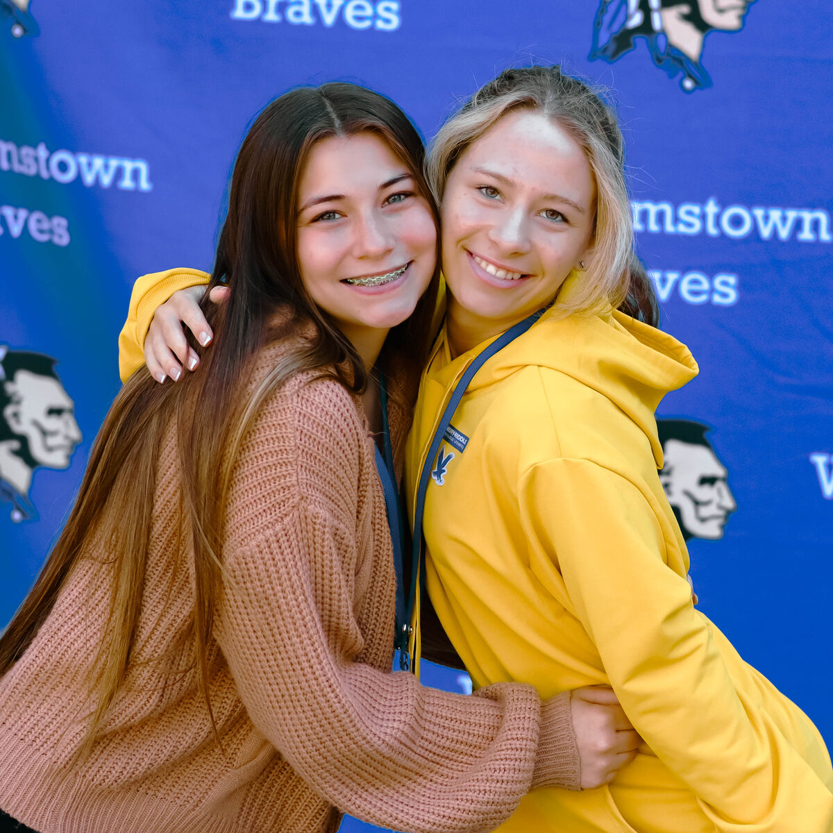 Two smiling young women embrace in front of a blue banner with text and graphics.