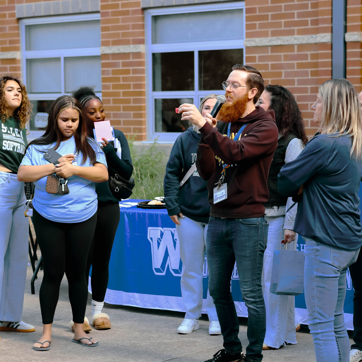 A man with a microphone speaks to a group of young people standing outdoors.