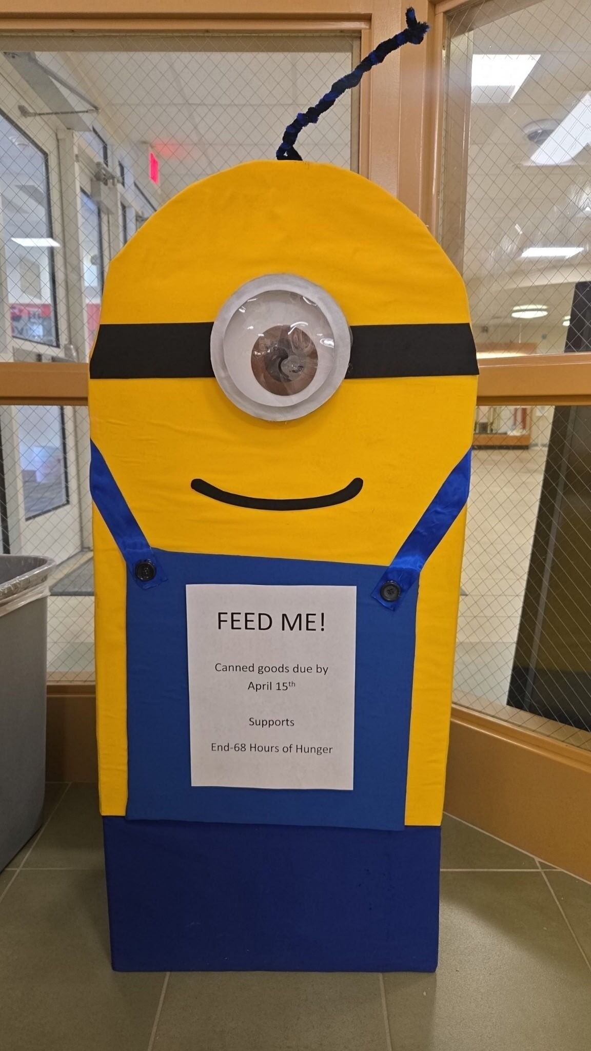 A yellow Minion-themed donation bin with a sign that reads 'FEED ME!' and details about canned goods.