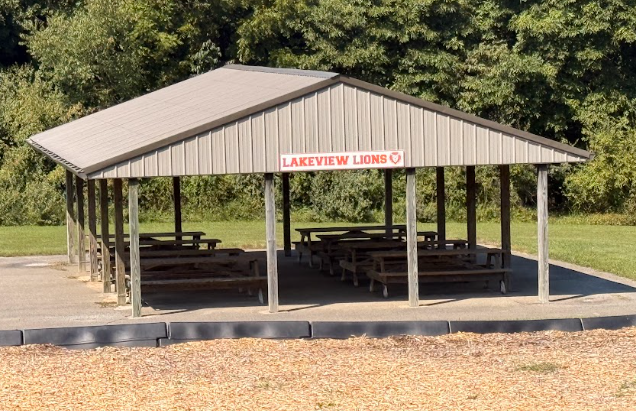 A covered pavilion with picnic tables sits in a grassy park surrounded by lush trees.