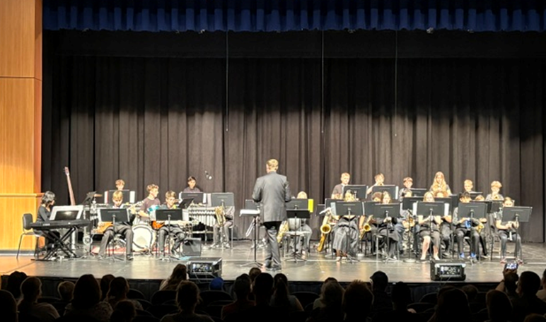 A school band performs on stage with a conductor facing the musicians.