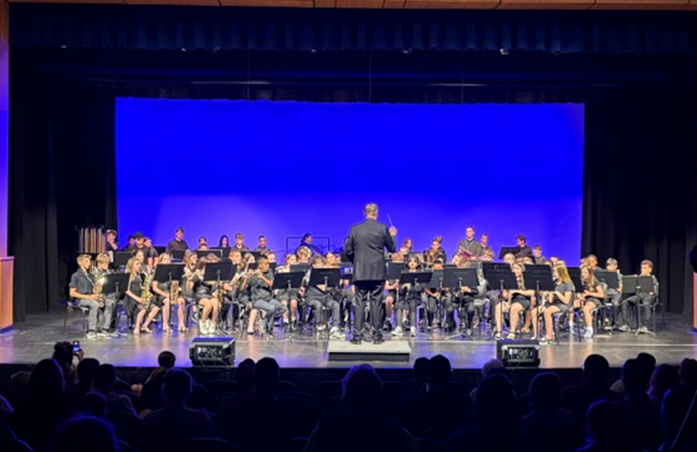 A conductor leads a large ensemble of young musicians playing various instruments on a stage.