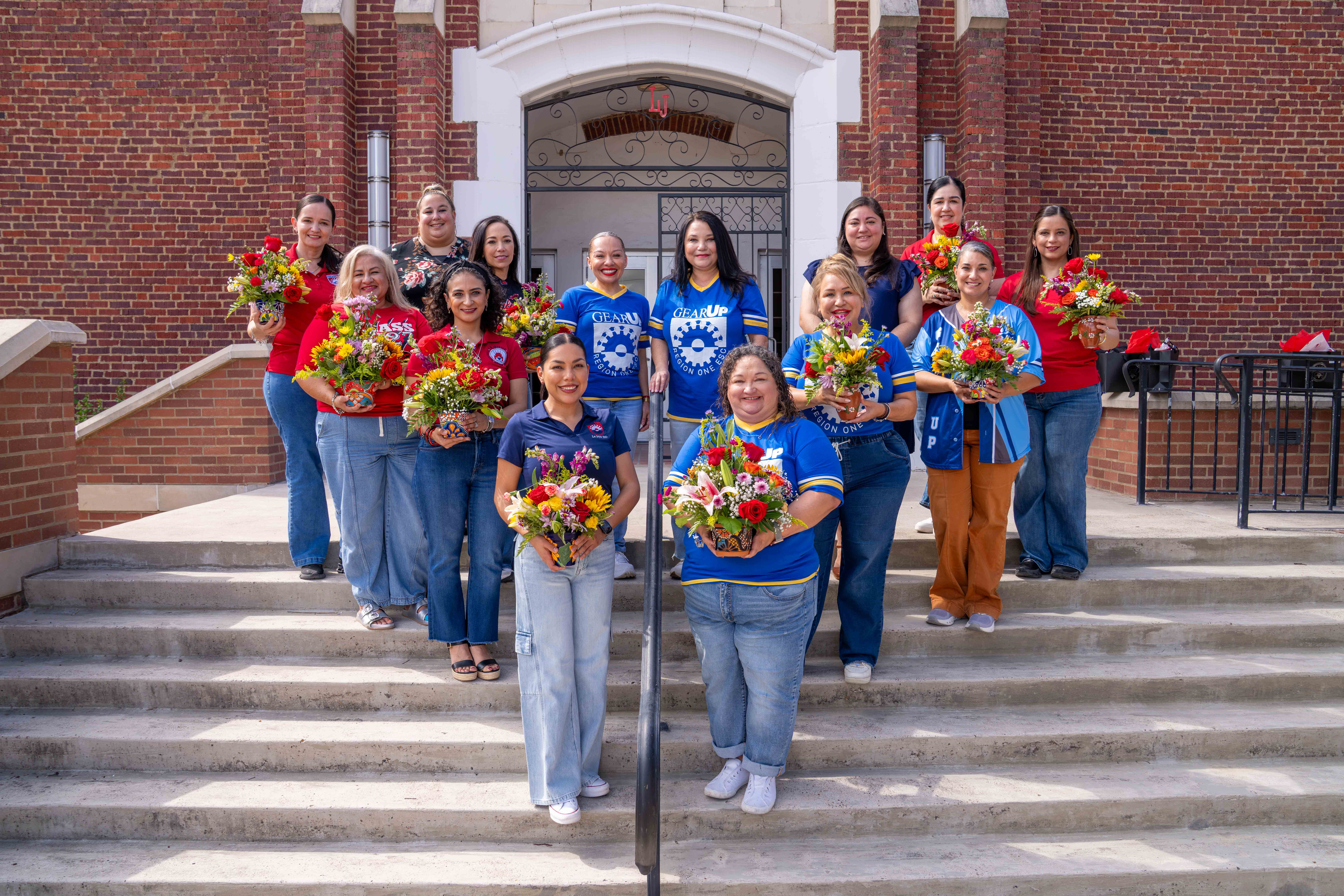 A group of women smiling while holding flower arrangements on the steps of a brick building with 'GEAR UP' shirts visible.