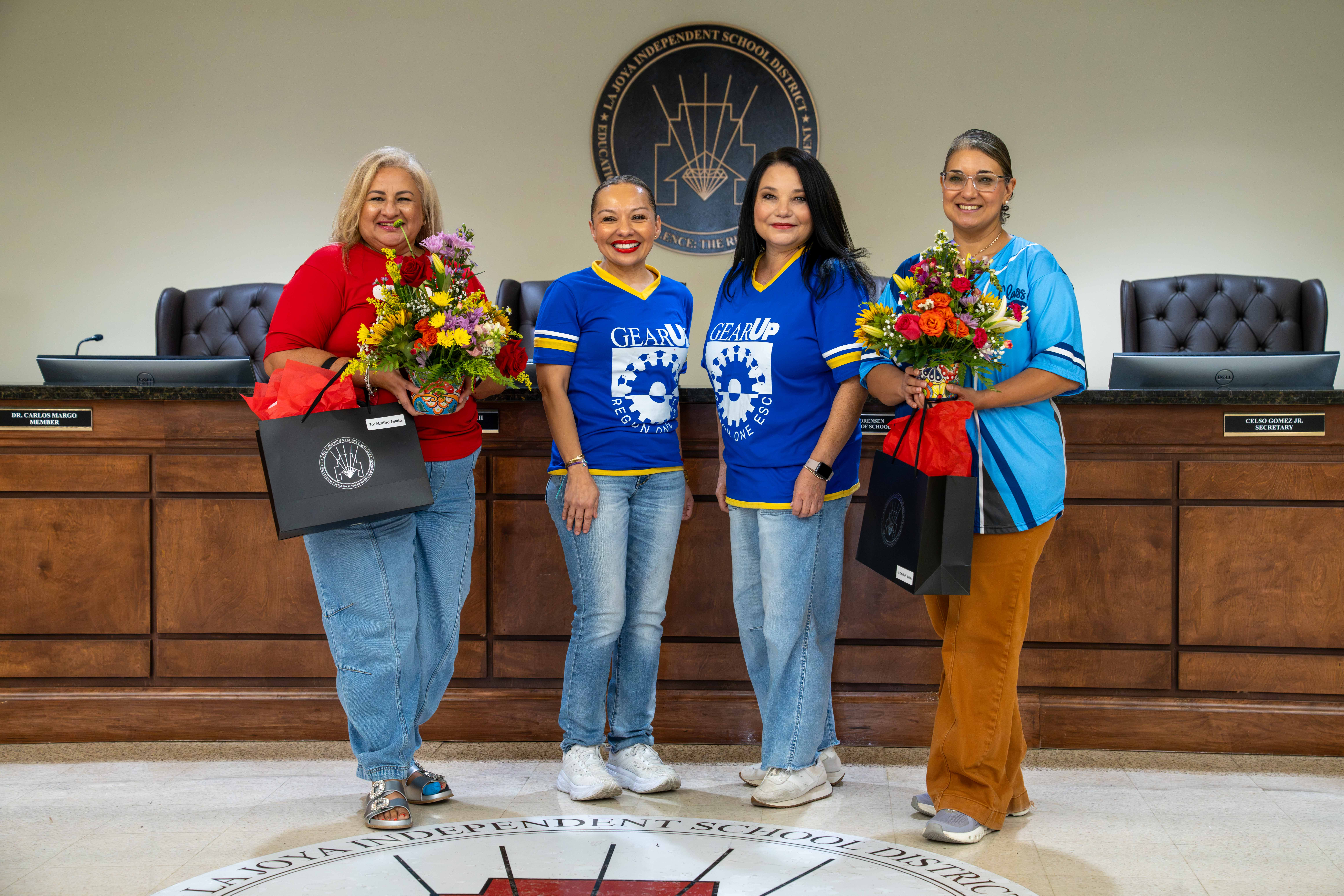 Four smiling women stand together in a school district board room, holding flower arrangements and gift bags.