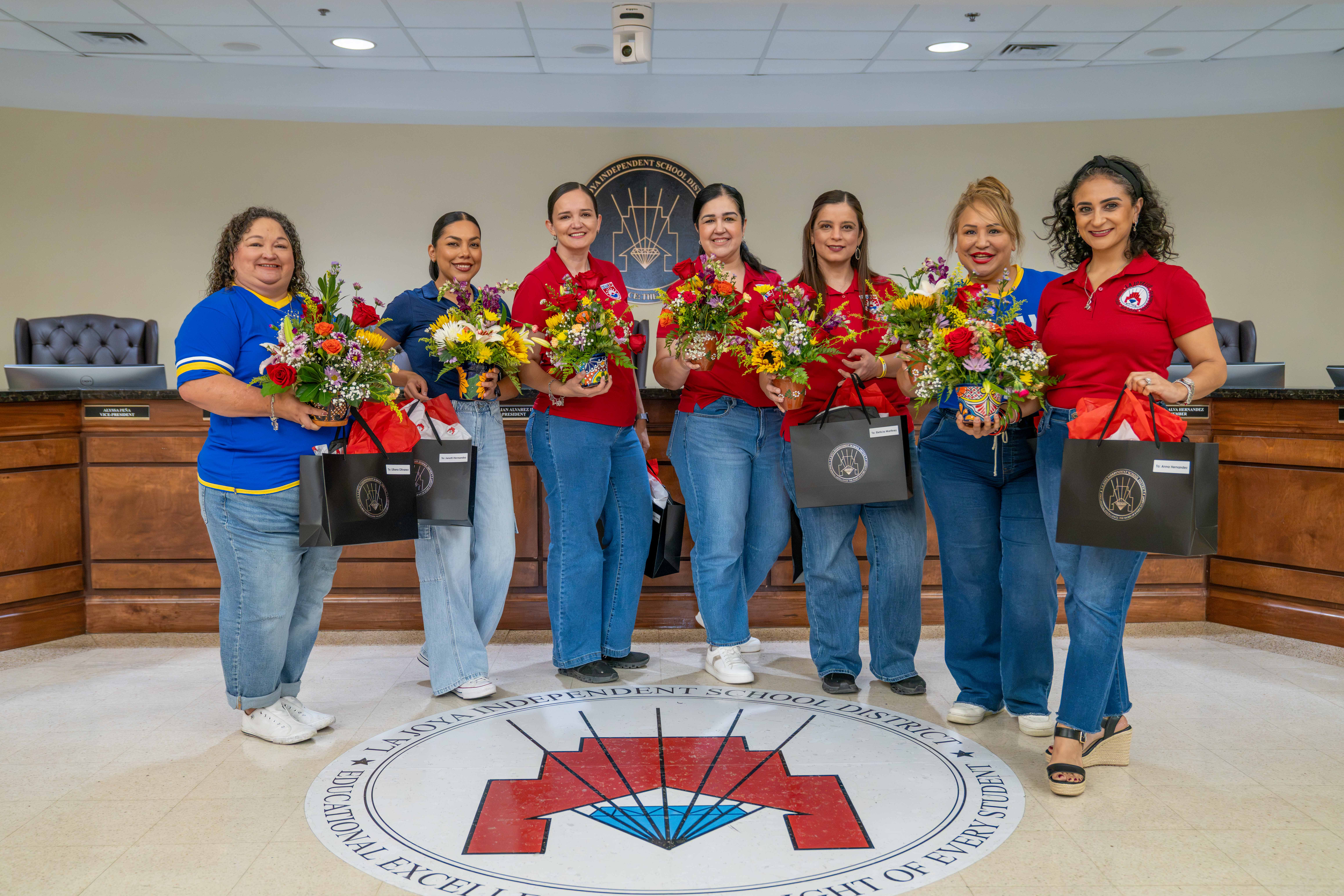 Seven smiling women stand in a semi-circle inside a meeting room, each holding a floral arrangement and a gift bag.