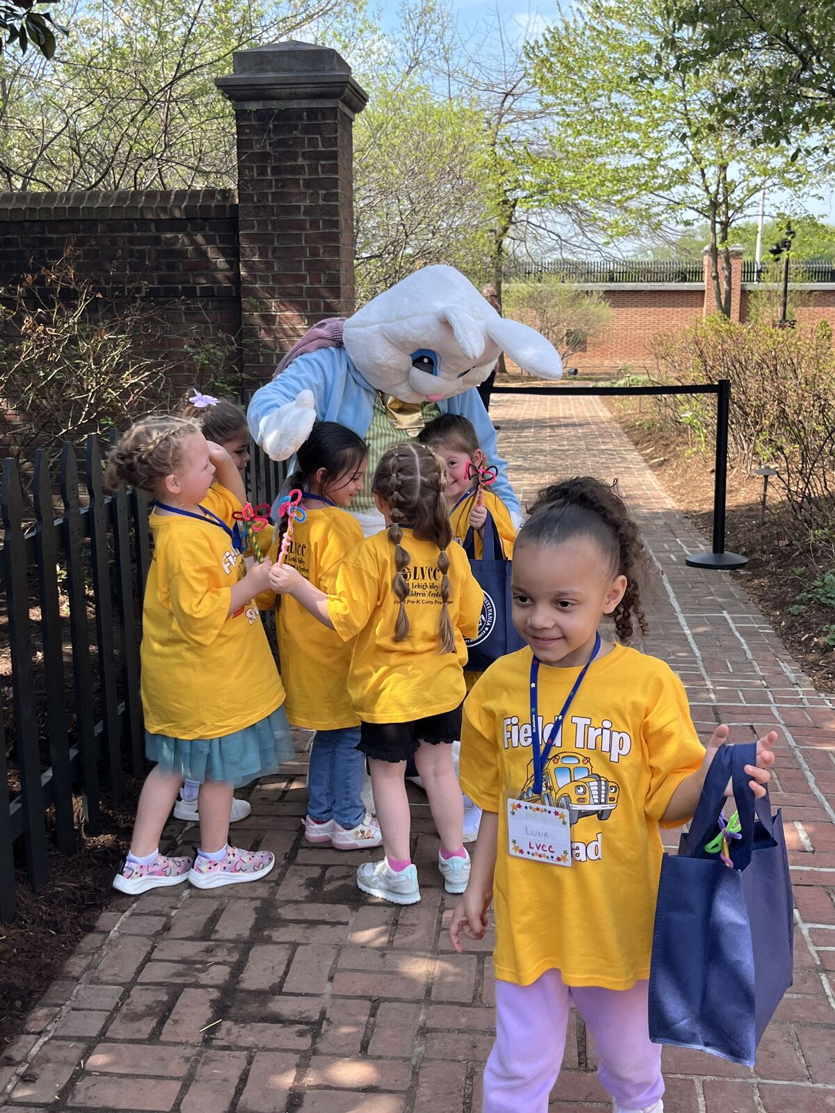 A group of children in yellow shirts stand with a person in a bunny costume on a brick pathway.