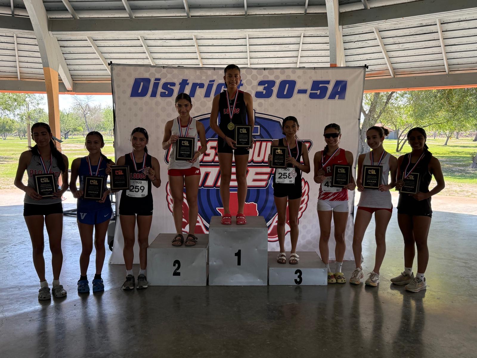 Eight young female athletes stand smiling on a podium, holding their awards and medals after a track meet.