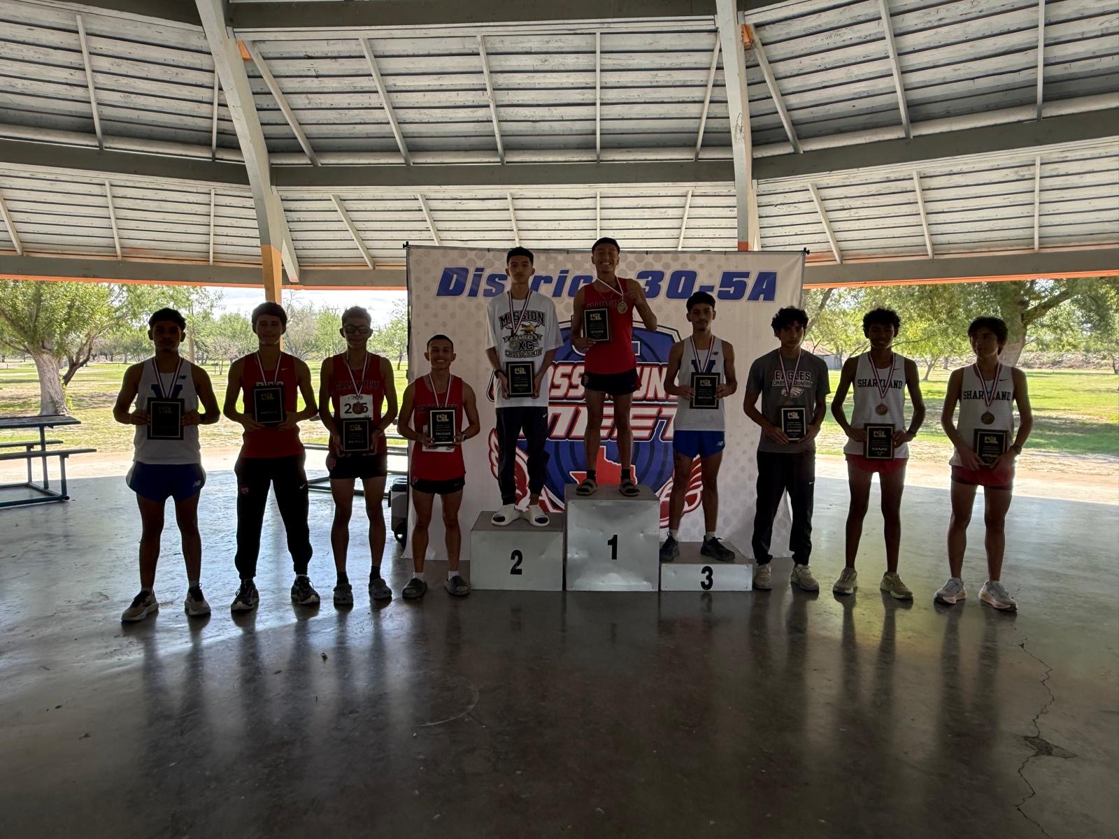 Ten young runners in athletic wear proudly pose together on and around a podium labeled "District 30-5A", holding plaques and medals.
