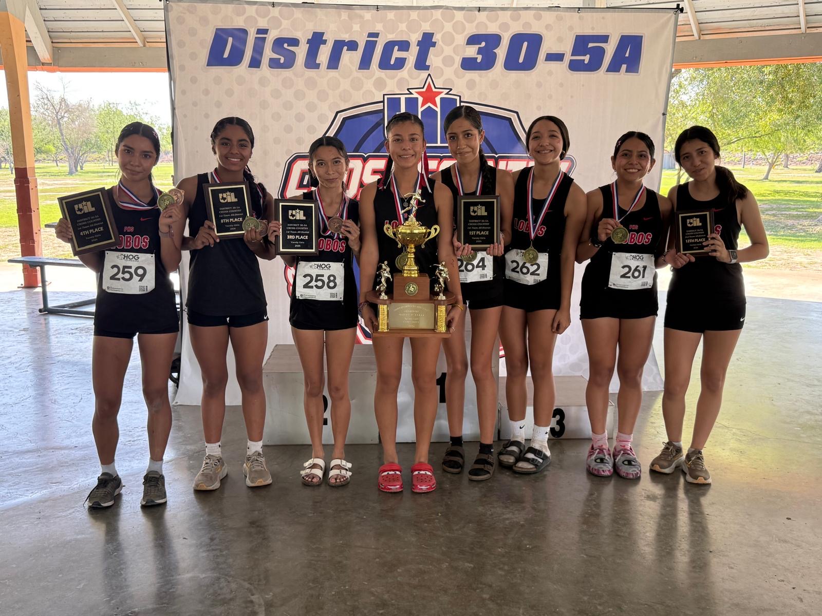 Seven female cross country runners in matching black uniforms proudly hold their awards and medals in front of a District 30-5A banner.