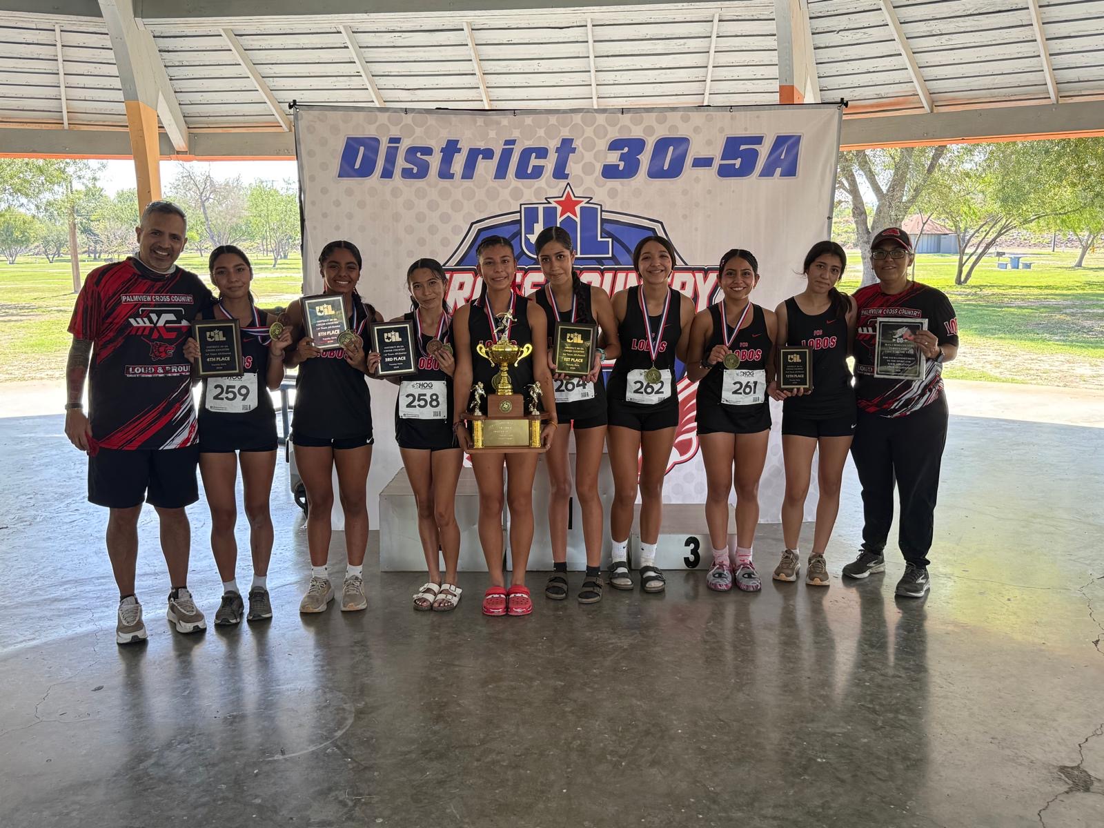 A cross country team holding trophies and medals poses proudly in front of a District 30-5A banner.