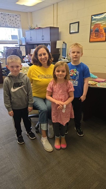 A smiling woman in a yellow shirt sits with three children in an office setting.