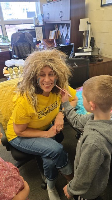 A child applies face paint to a smiling woman wearing a blonde wig and a yellow t-shirt.