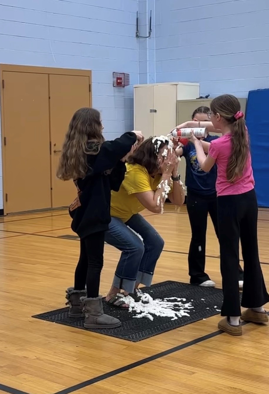 Children playfully spray whipped cream on a person's face in a gymnasium.