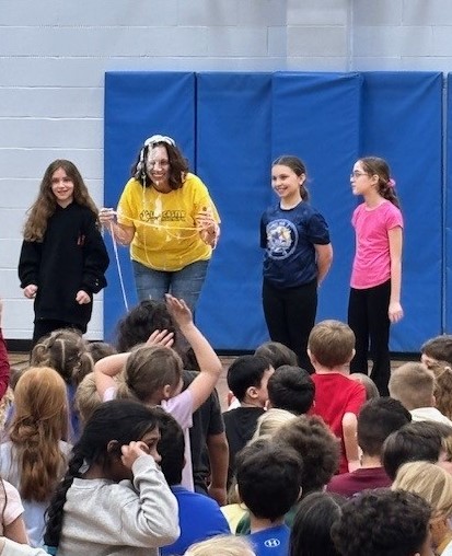 A teacher with whipped cream on her face smiles at a crowd of students in a gymnasium.