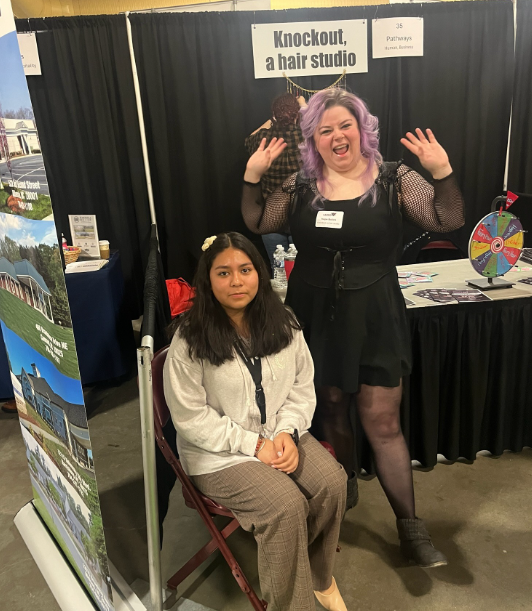Two women stand at a booth for "Knockout, a hair studio" with a prize wheel in the background.