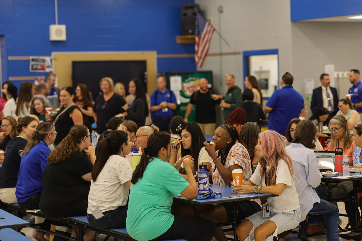 A group of people are gathered around tables in what appears to be a school cafeteria, some eating and conversing.