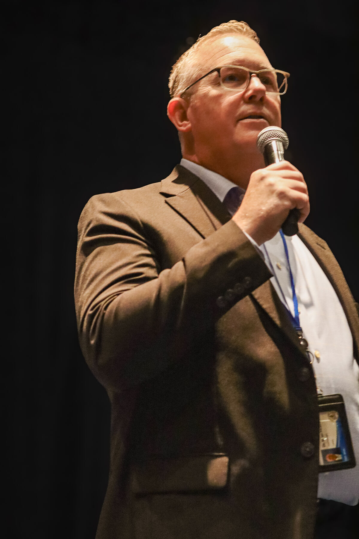 A man in a suit and glasses speaks into a microphone on a dark stage.