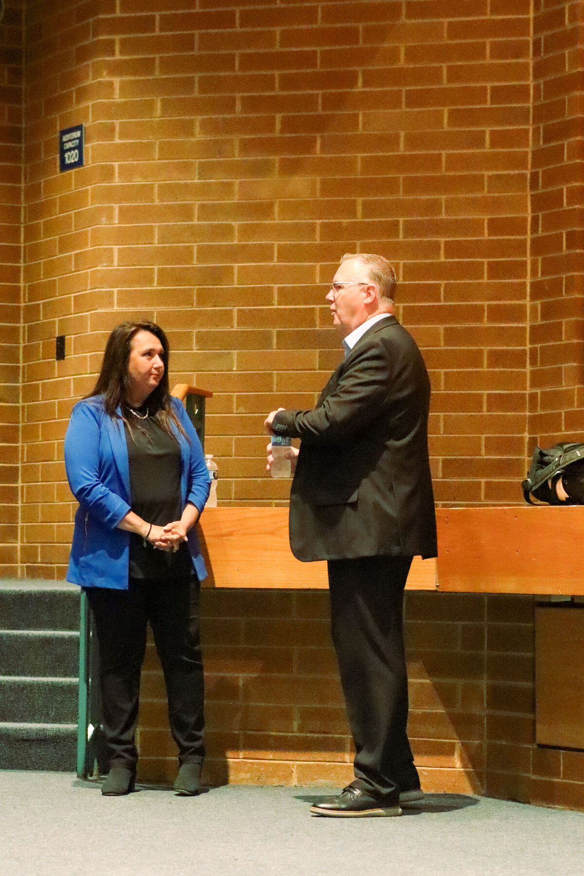 A woman in a blue blazer and a man in a suit stand and talk in front of a brick wall.
