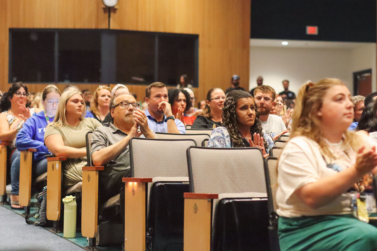 An audience sits attentively in a lecture hall with wooden walls and a large screen.