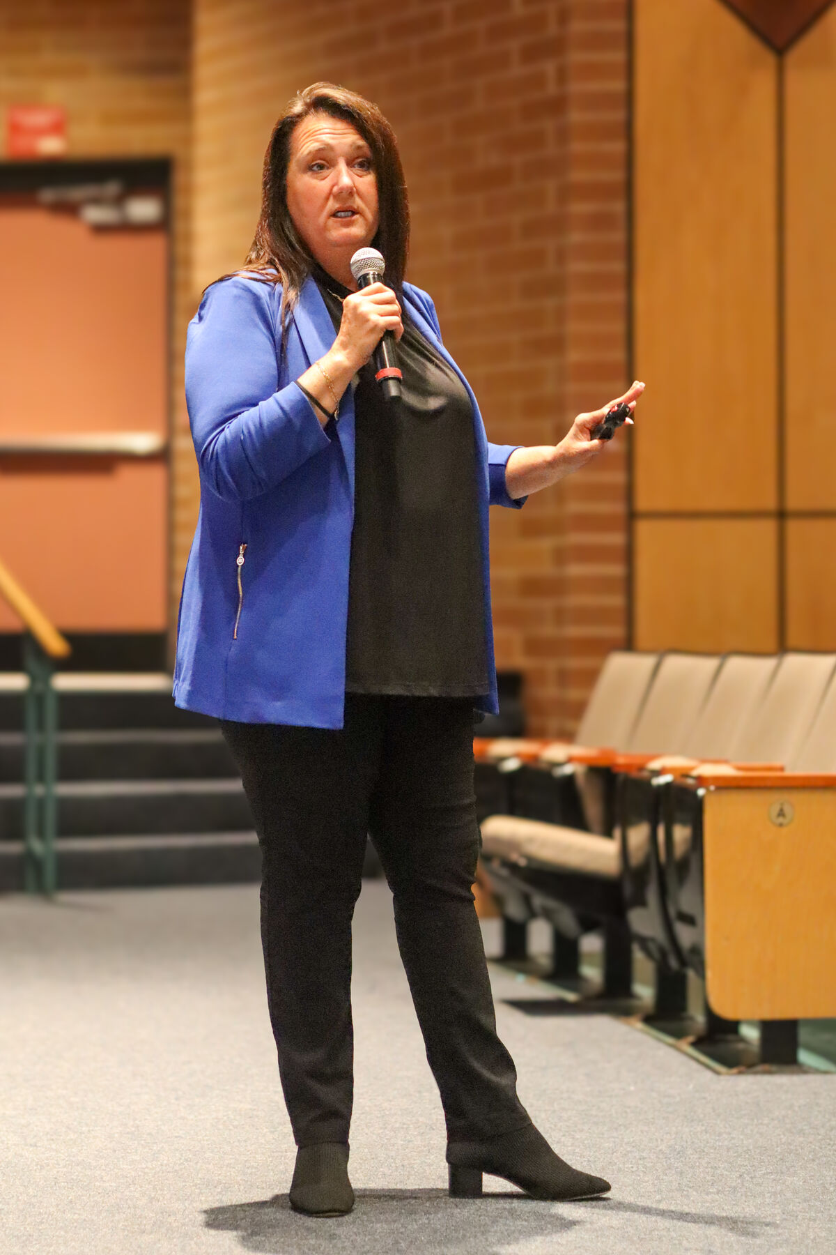 A woman in a blue blazer speaks into a microphone while holding a clicker in an auditorium.