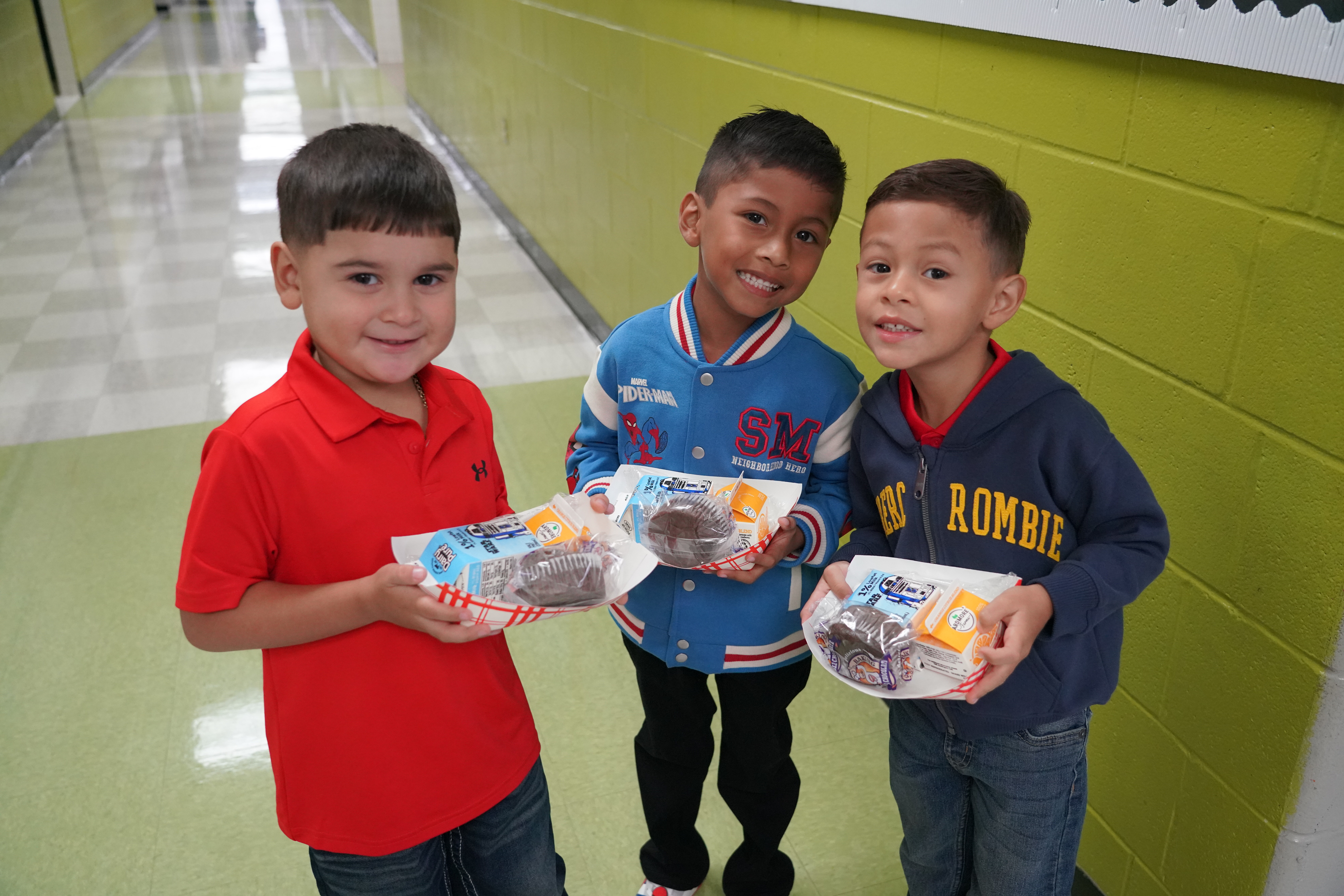 Three smiling boys stand in a school hallway, each holding a tray with a muffin, juice box, and milk.