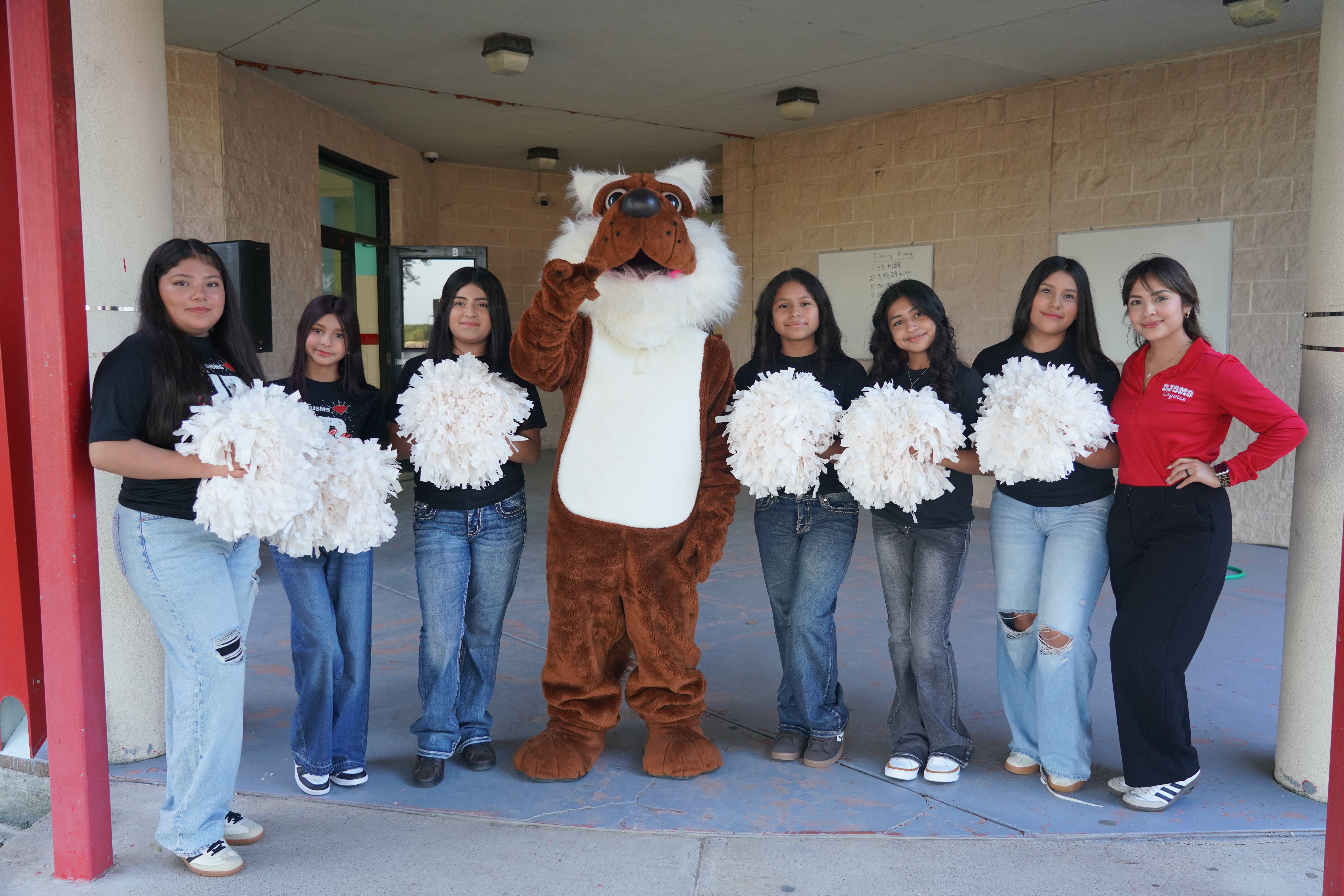 A group of students and a mascot stand together outside a building, holding white pom-poms.