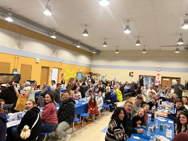A large group of people, including children and adults, are seated at long tables in a brightly lit room, appearing to be at an event or gathering.