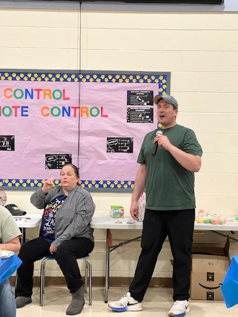 A man in a green shirt and cap speaks into a microphone, standing near a bulletin board with text about 'REMOTE CONTROL'.