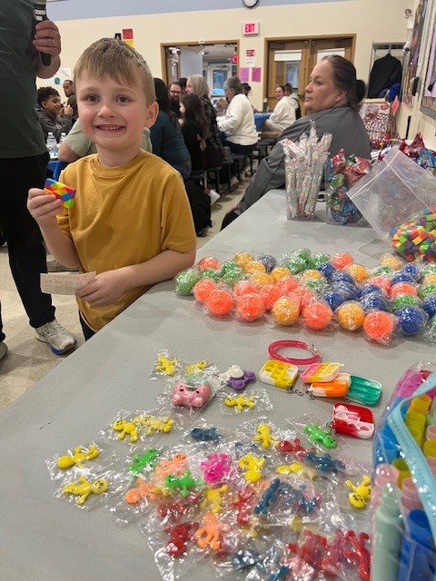 A young boy smiles, holding a colorful fidget toy at a table filled with small toys and prizes.