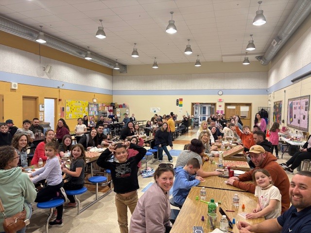 A large group of families are gathered at long tables in a school cafeteria, enjoying an event.