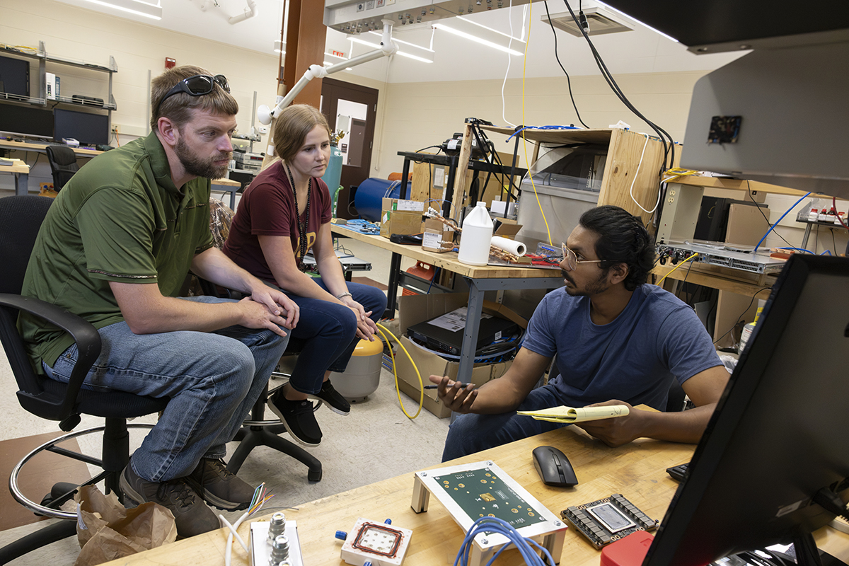 Three people collaborate in a lab, discussing electronics and equipment.
