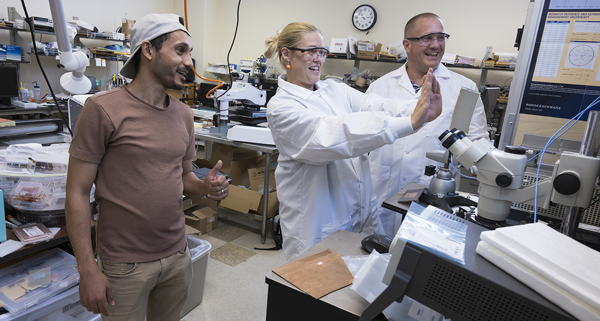Three people in a laboratory setting, two in lab coats, interact near scientific equipment.
