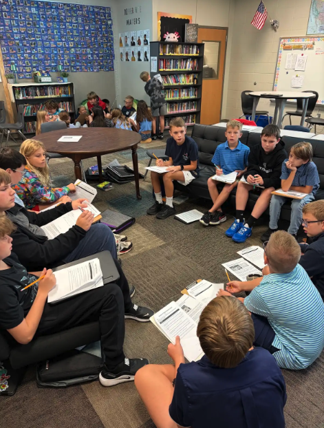 A group of students sit in a circle on the carpet of a classroom, reading papers with pencils.