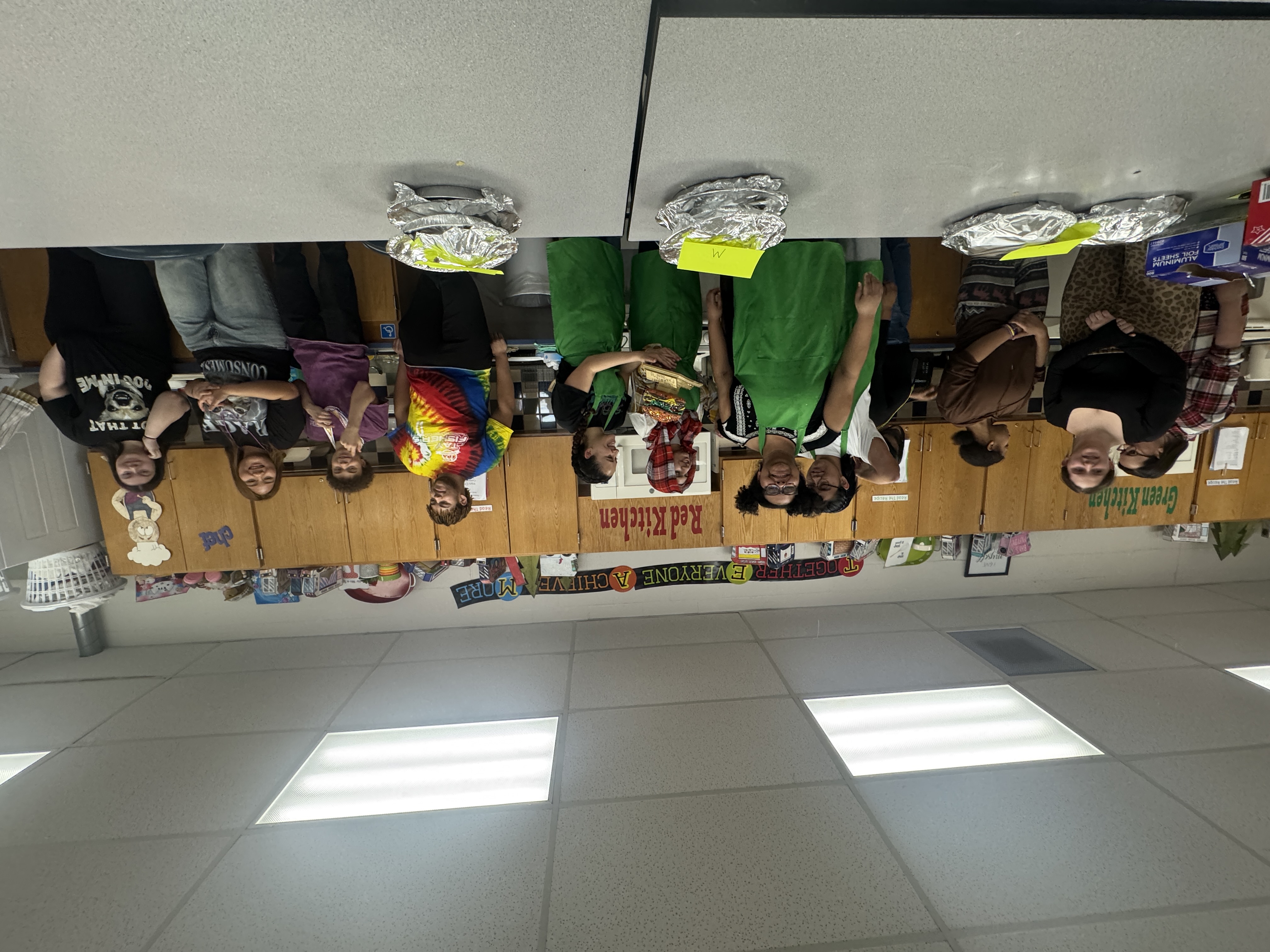 A group of students stand behind a table with foil-wrapped dishes and green aprons.